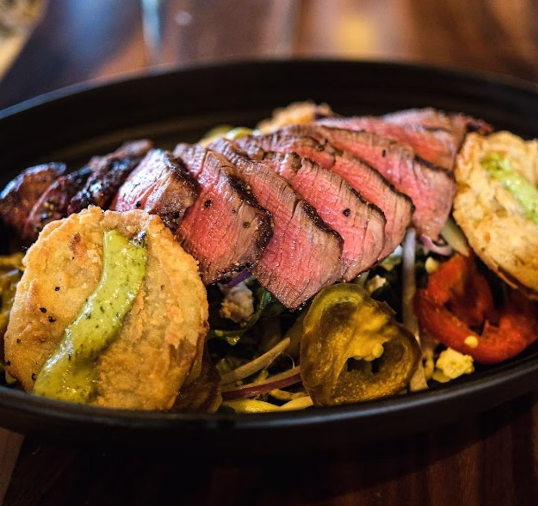 Sliced steak, fried discs with green sauce, peppers, and greens.