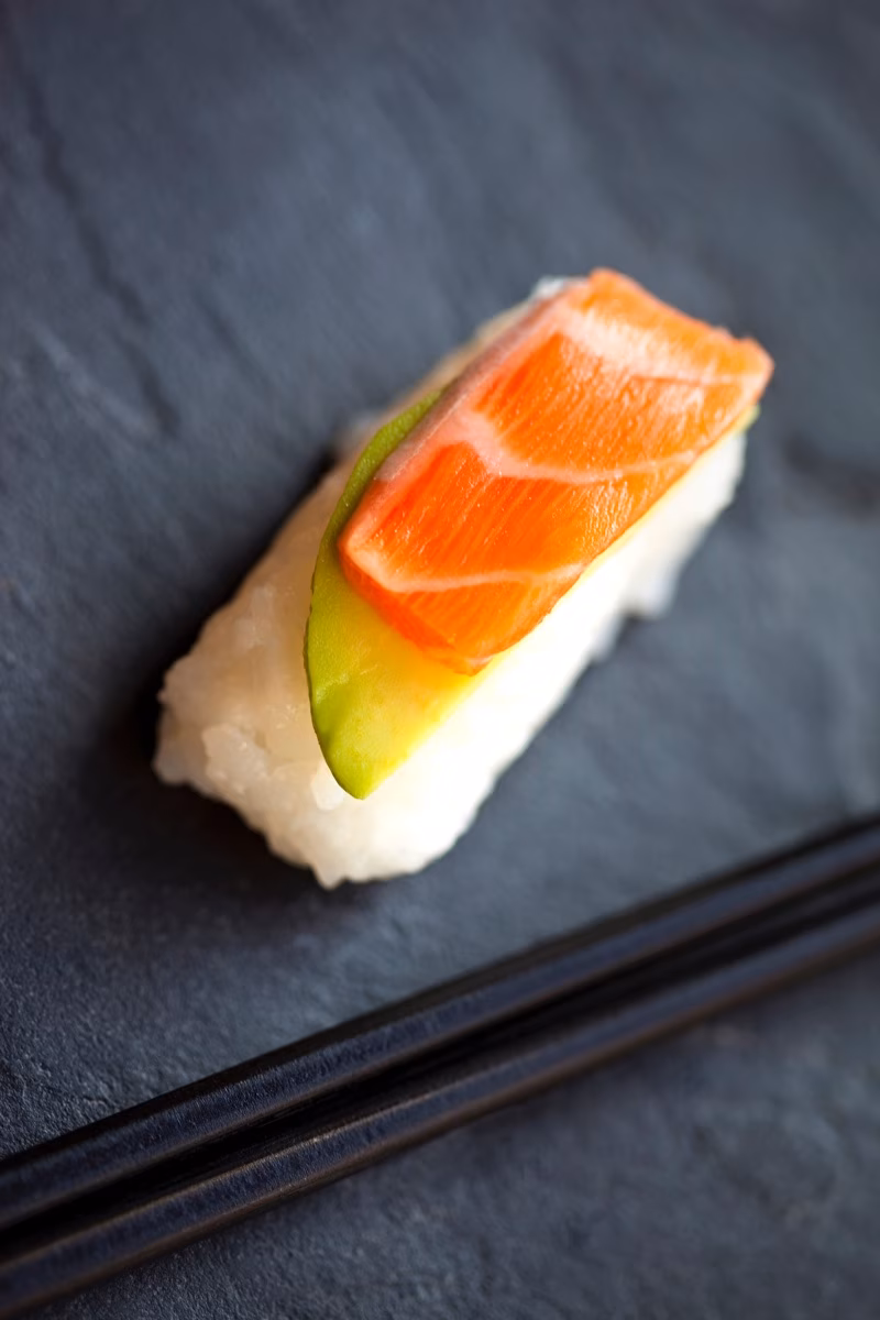 Single salmon and avocado nigiri sushi with chopsticks on slate.