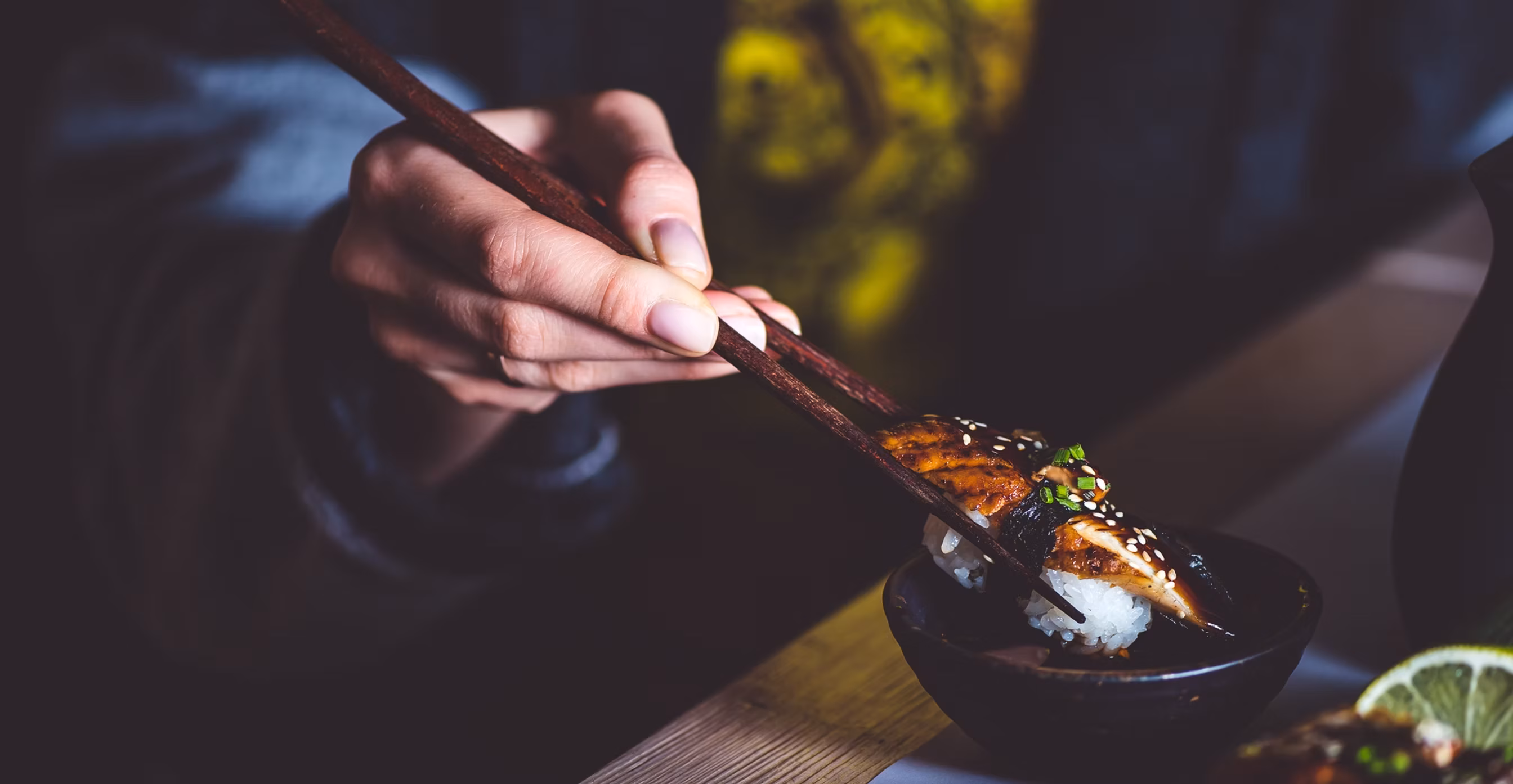 Hand using chopsticks to pick up eel sushi from a dark bowl.