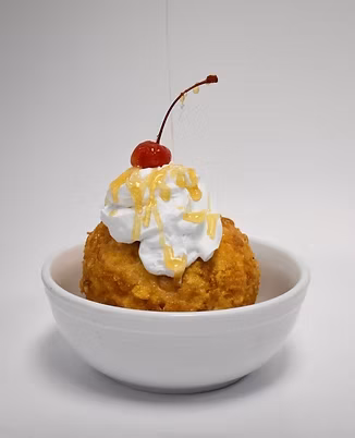 Deep-fried dessert ball with whipped cream, honey, and cherry in a bowl.