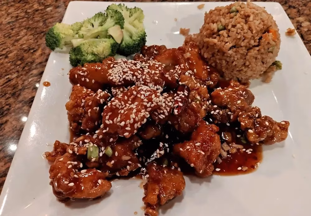 Sesame chicken with fried rice and broccoli on a white plate.