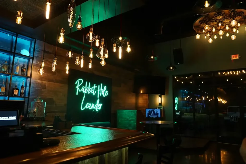Dimly lit bar interior with glowing counter and "Rabbit Hole Leander" neon sign.