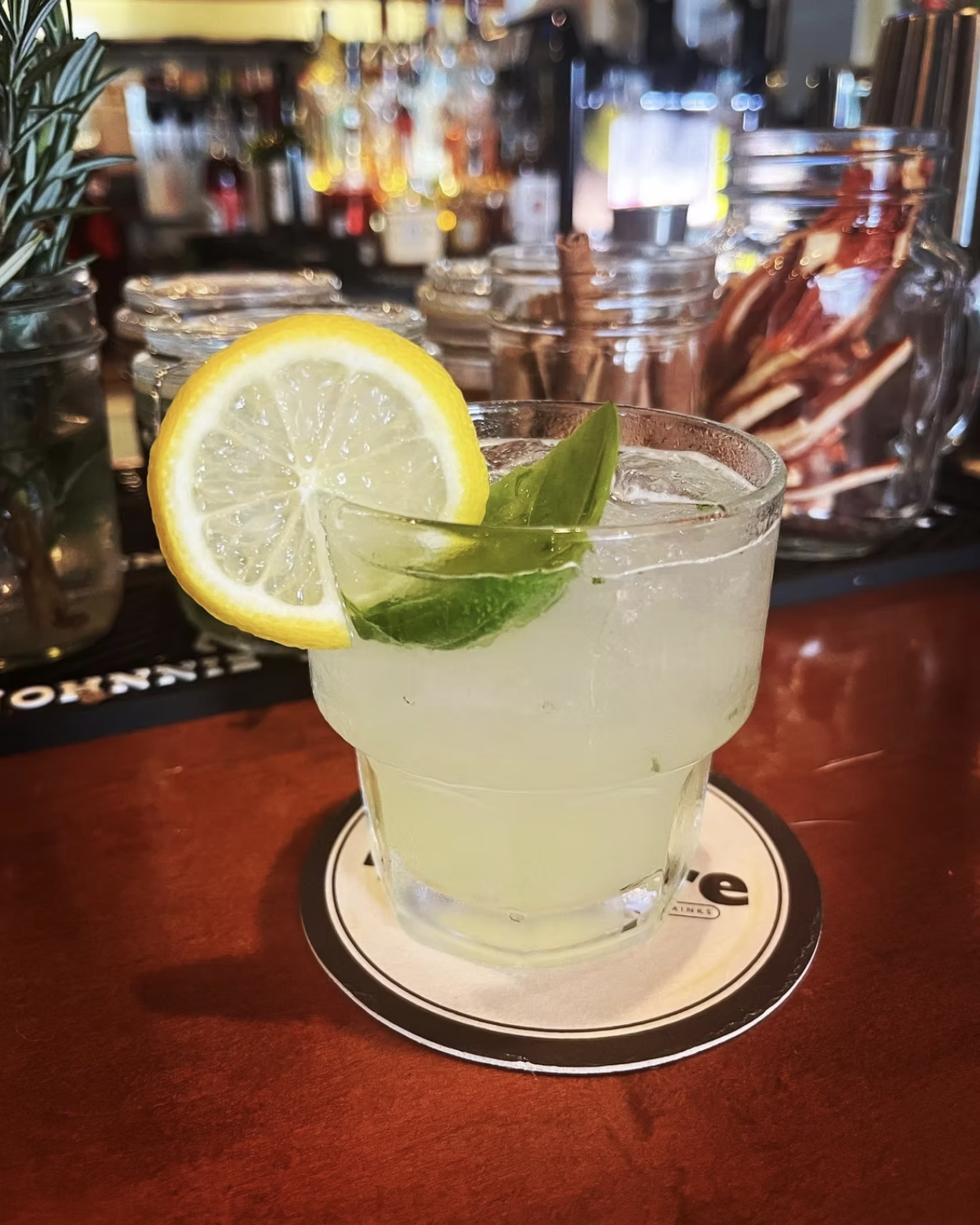 An iced cocktail with a lemon slice and green leaf on a bar.