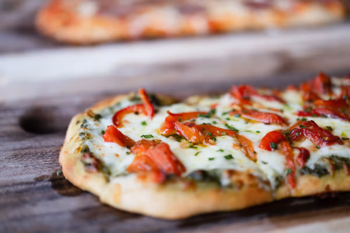 Flatbread with melted cheese, red peppers, and green herbs on wood.