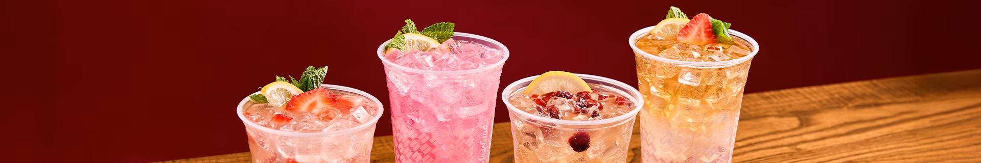 Four iced drinks with fruit and mint garnishes on a wooden surface.