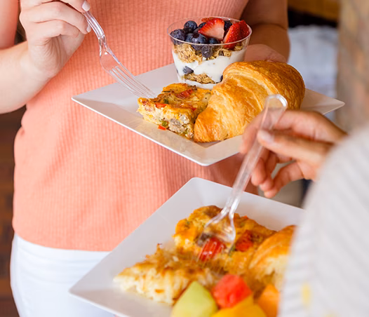 Hands holding plates of breakfast quiche, croissant, fruit, yogurt parfait.