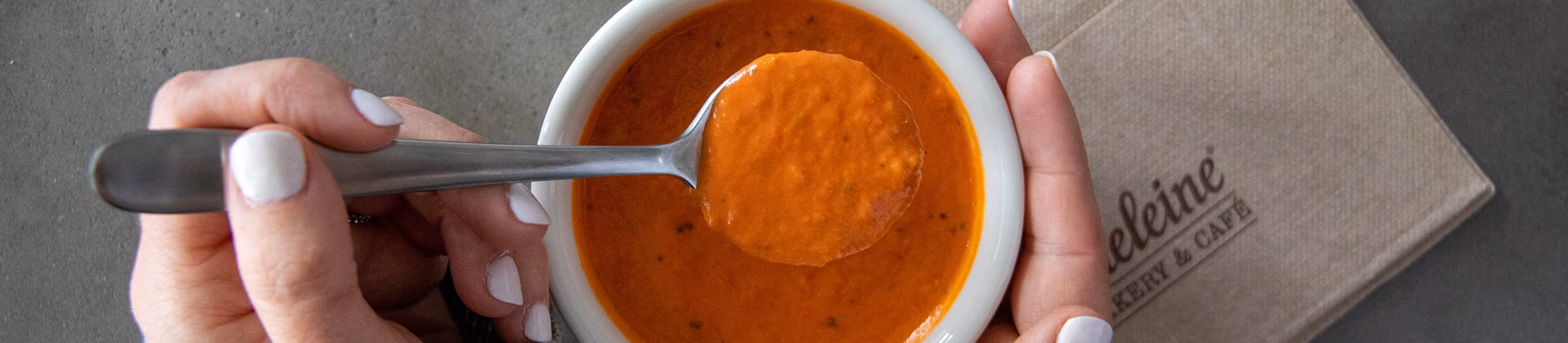 Hands scooping orange soup from a white bowl with a spoon.
