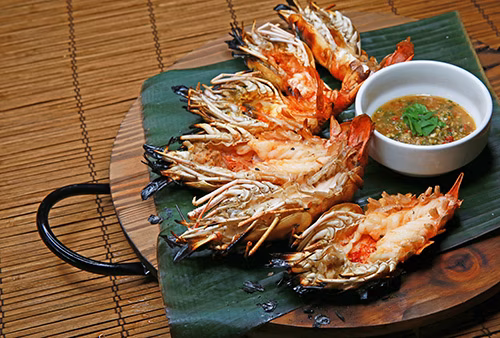 Grilled split prawns with dipping sauce on a wooden platter.