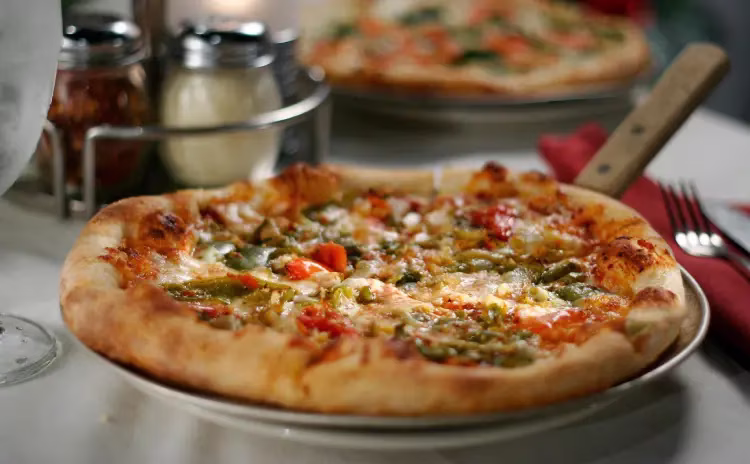 A pizza with green and red toppings on a table with another pizza.