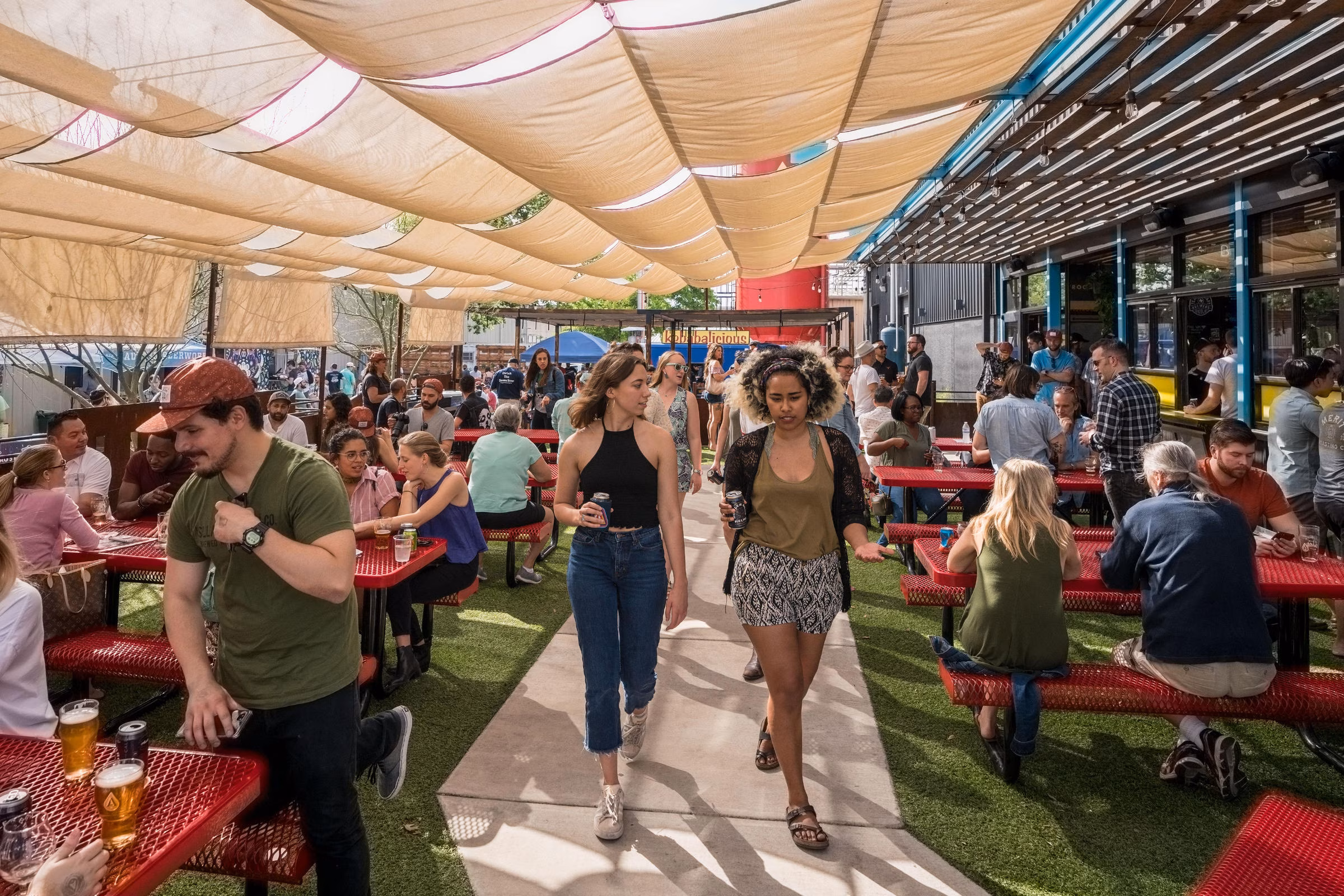People gathered at an outdoor space with red picnic tables and shade.