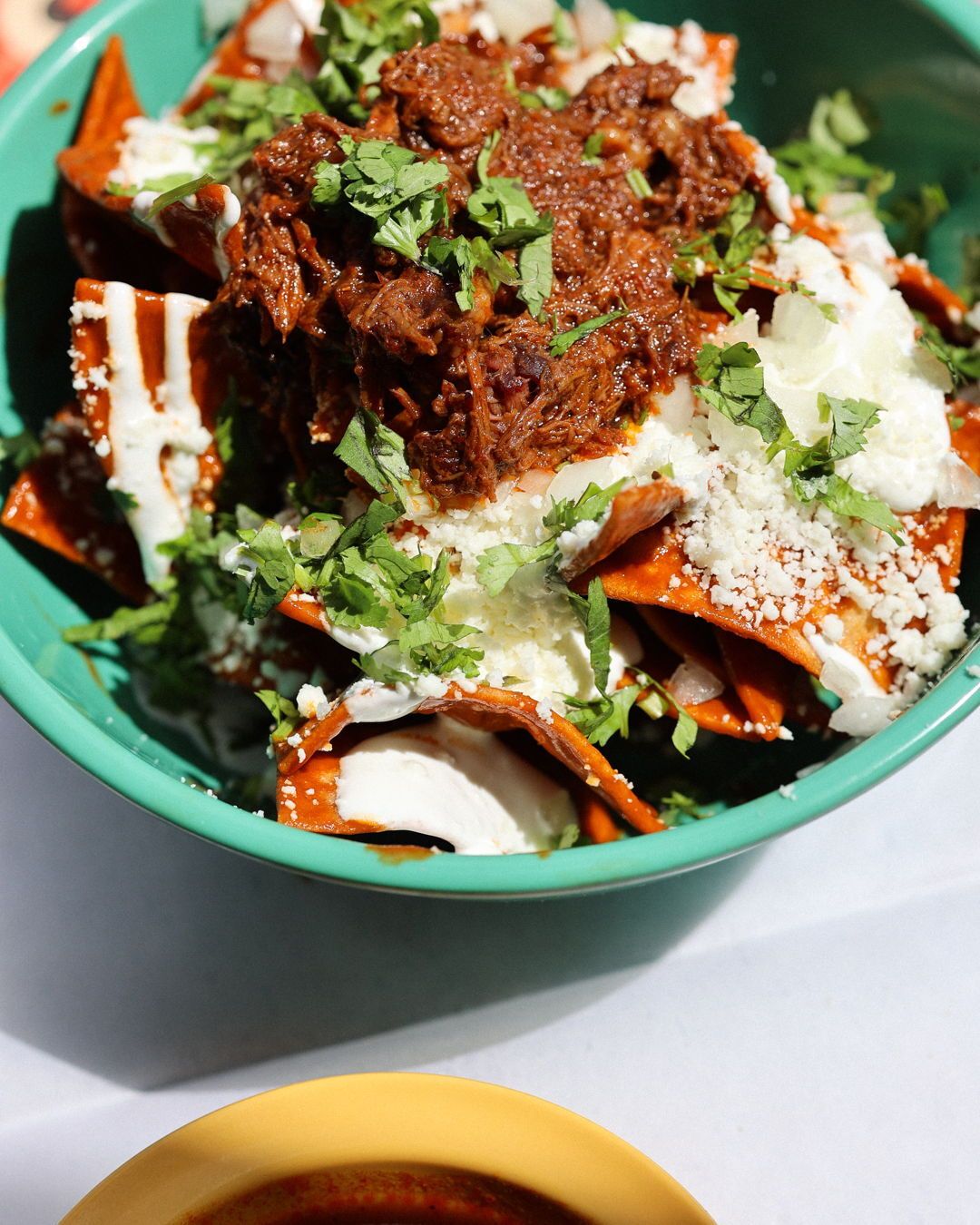 Chilaquiles with shredded meat, cheese, crema, and cilantro in a teal bowl.