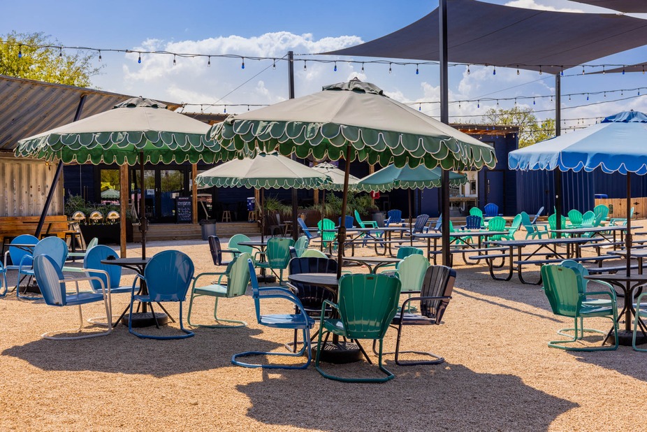 Gravel patio with blue and green chairs under umbrellas and shade.