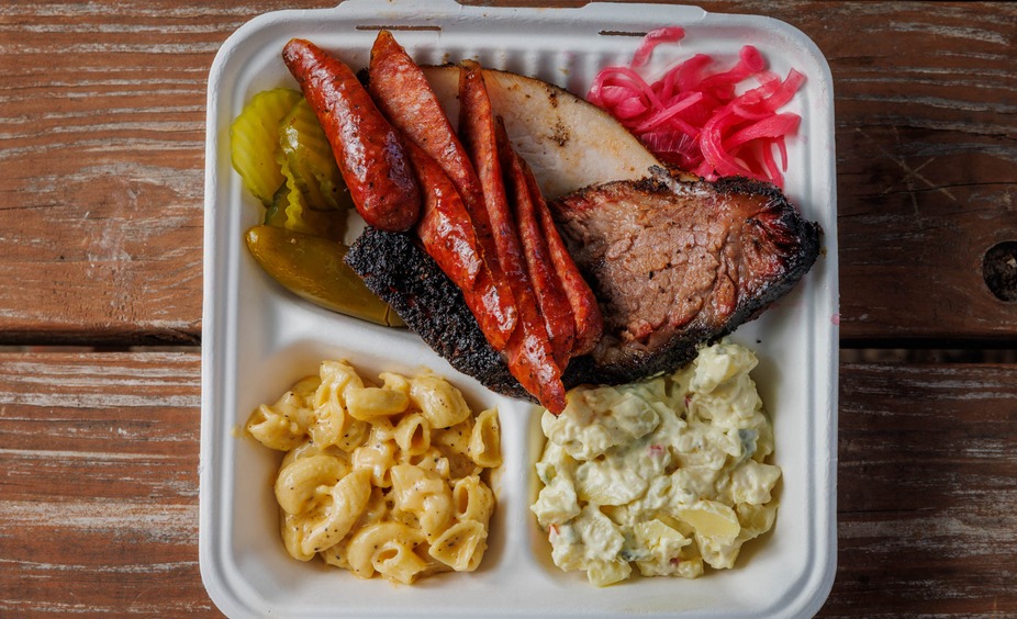 Takeout container with smoked meats, macaroni, potato salad, and pickles.