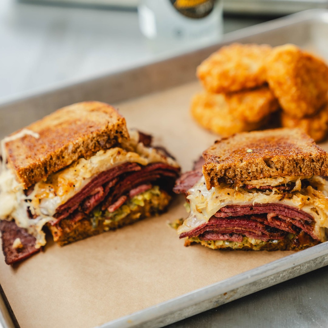 Two Reuben sandwiches and fried hash browns on a tray.