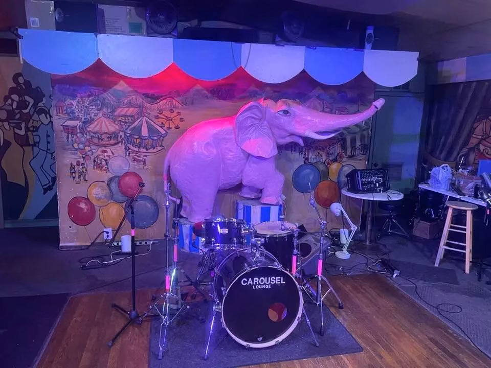 Pink elephant statue, drum set, and carnival mural on stage.