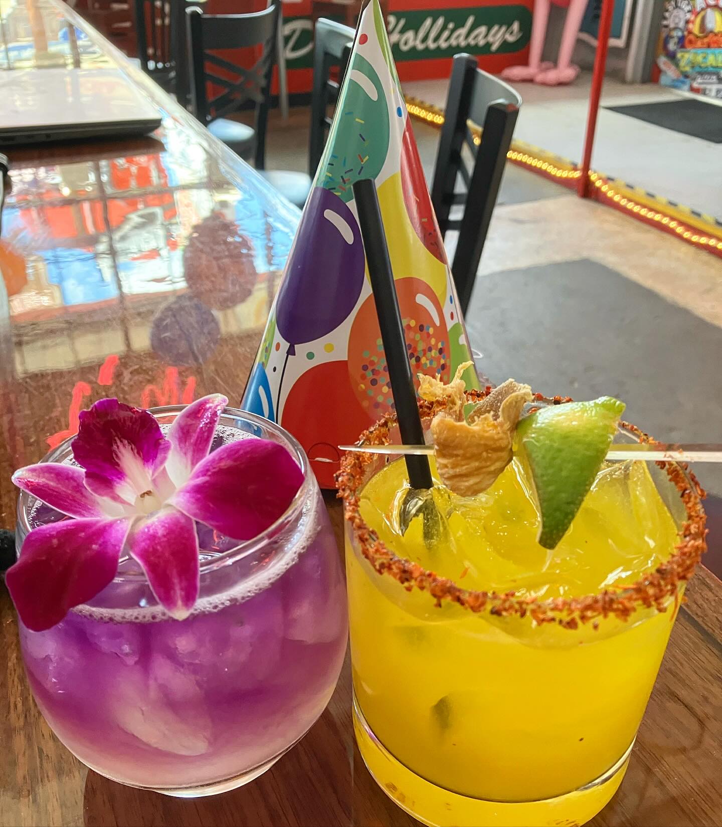 Purple and yellow cocktails, a birthday hat, on a wooden bar.