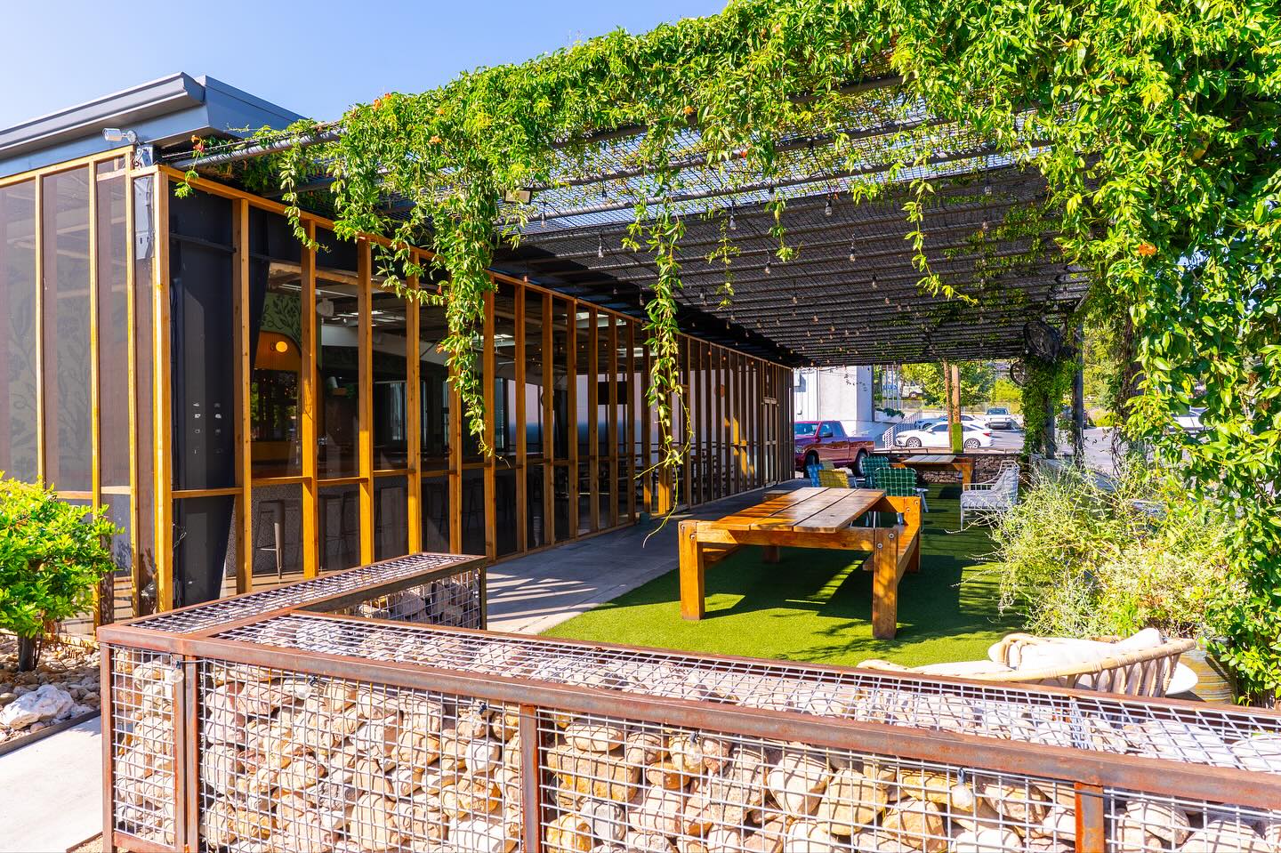 Outdoor patio with vine-covered pergola, wooden tables, and gabion wall.