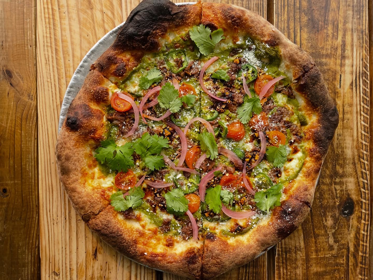 Pizza with green sauce, crumbled meat, tomatoes, red onions, and cilantro.