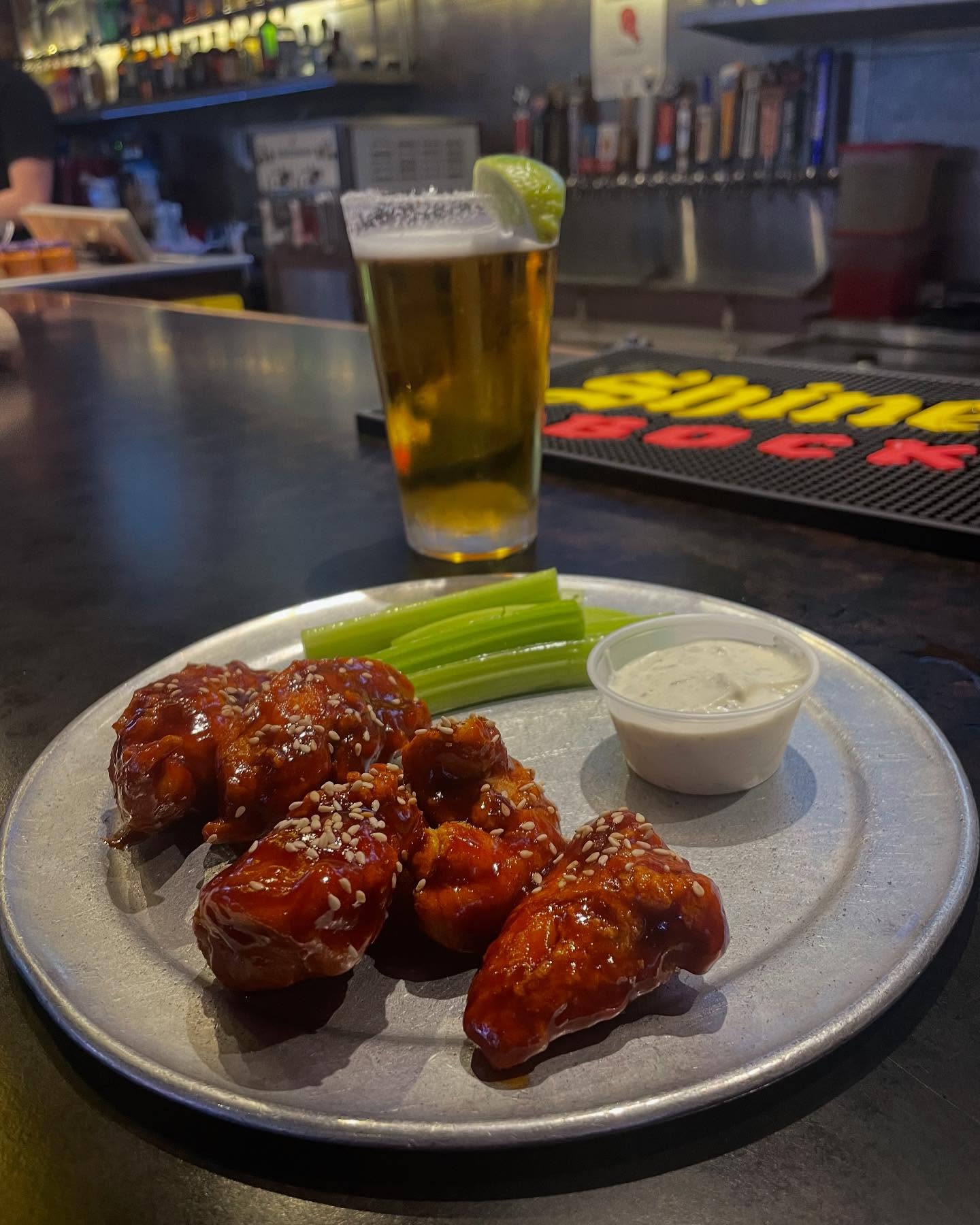 Glazed chicken wings, celery, dipping sauce, and beer on a bar counter.