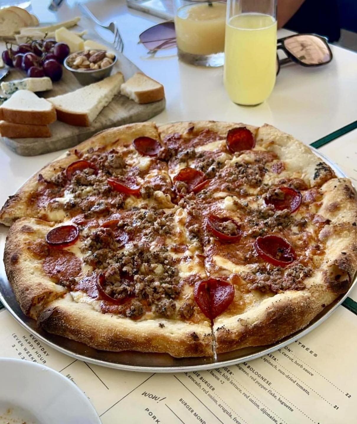Pepperoni and sausage pizza, charcuterie board, and drinks on white table.