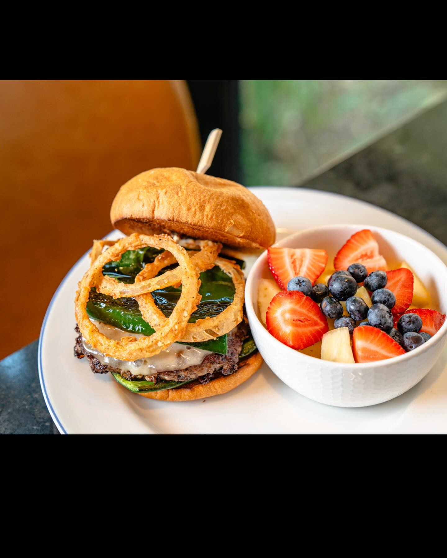 Burger with onion rings, cheese, peppers, and a side of fruit salad.