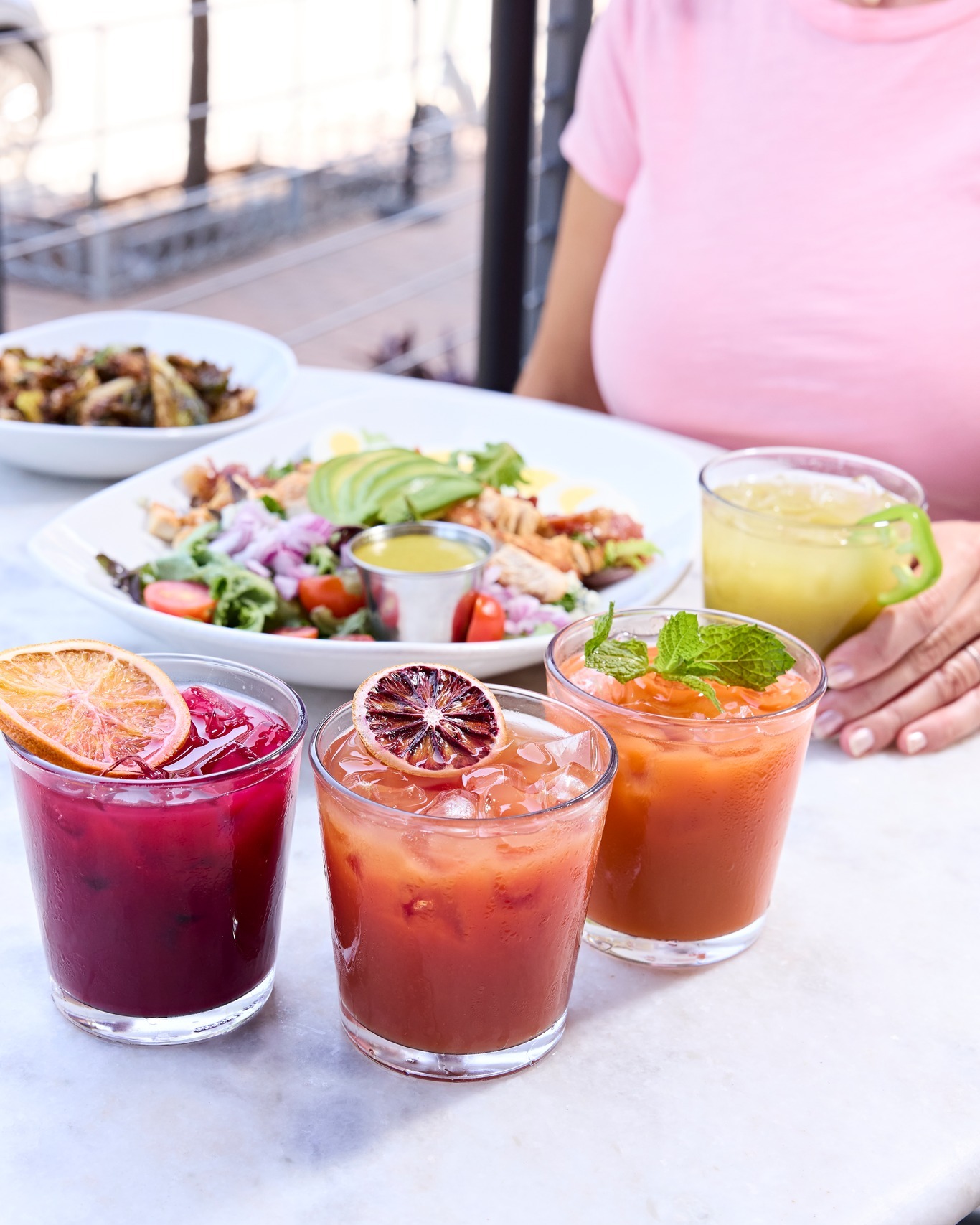 Three colorful cocktails, salad, and a side dish on a marble table.
