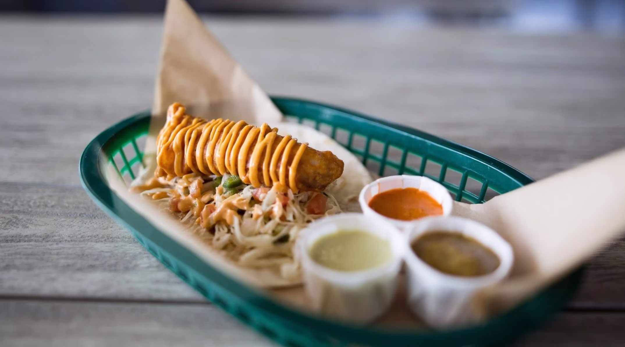 Taco with battered item, spiral sauce, three dips, in green basket.