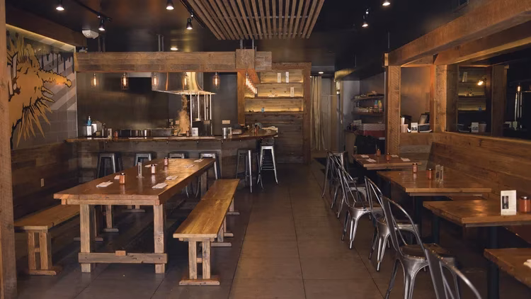 Restaurant interior with wooden tables, benches, a bar, and metal chairs.