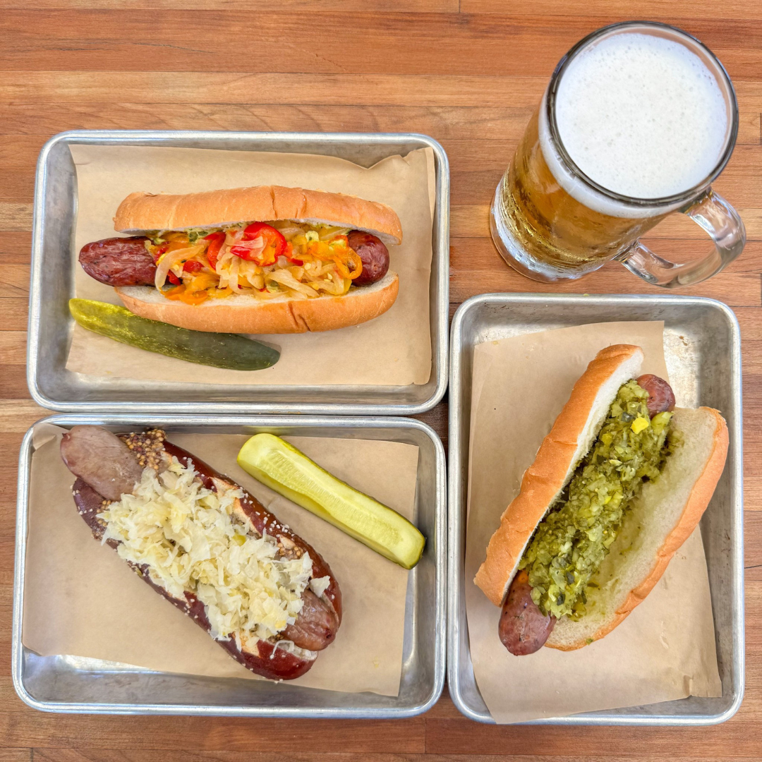 Three sausages with assorted toppings, pickles, and a beer.