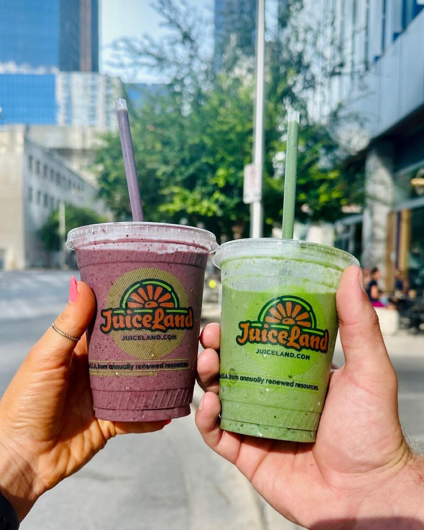 Two hands holding purple and green JuiceLand smoothies outdoors.