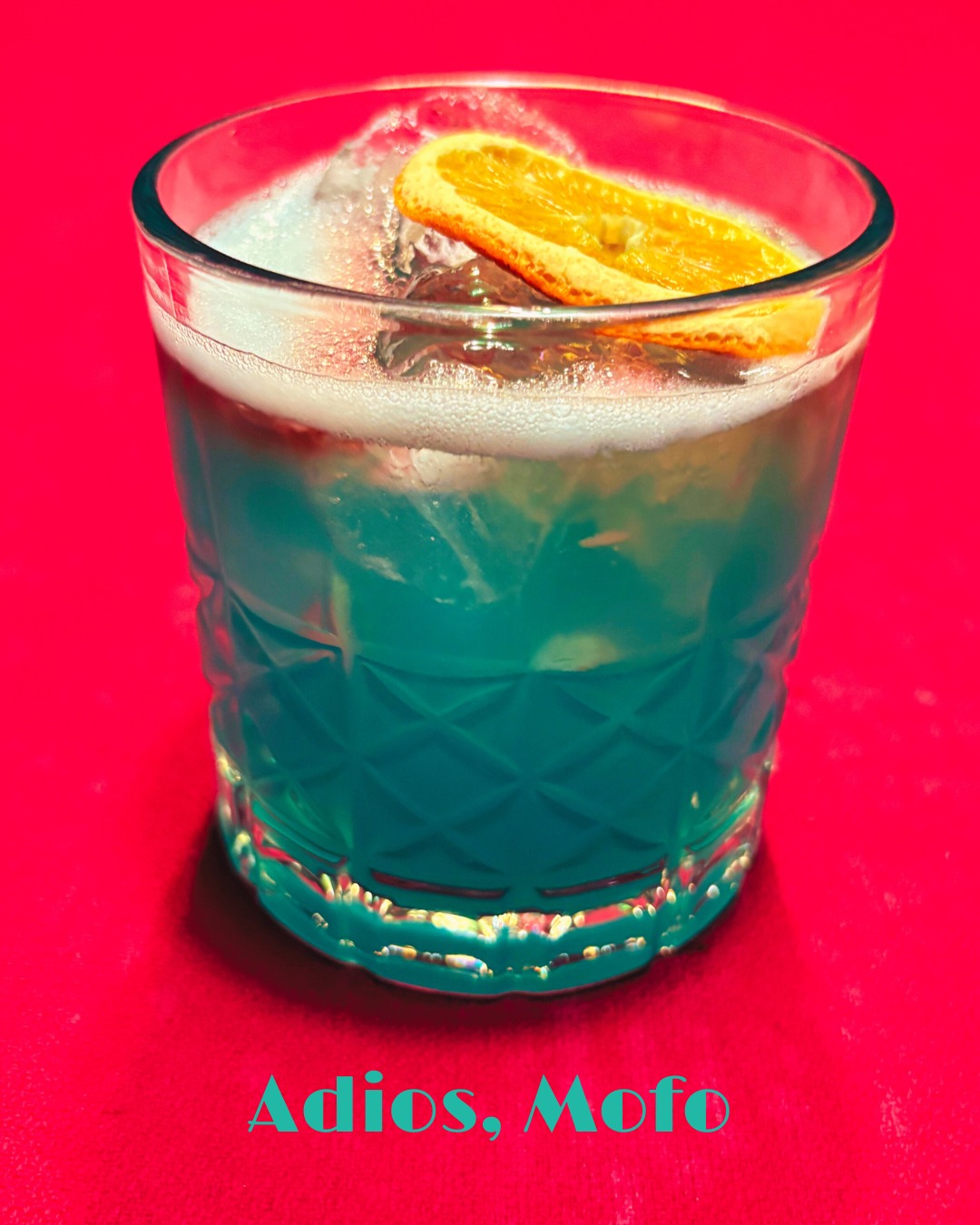Blue-green drink with ice, foam, and orange slice on red.