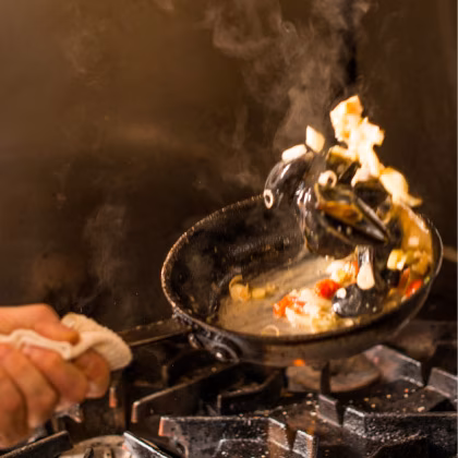 Chef tossing mussels and vegetables in a steaming pan on a stovetop.
