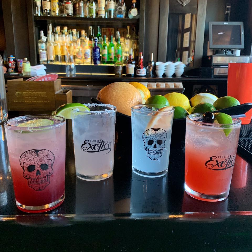 Four colorful cocktails in skull-decorated glasses with lime on a bar.