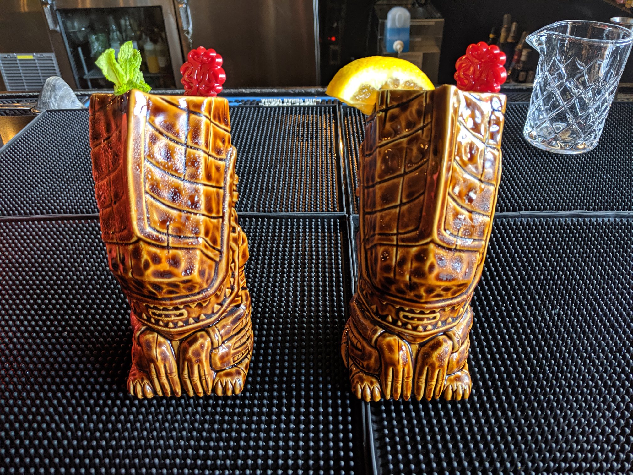 Two turtle-shaped tiki mugs with garnished cocktails on a bar mat.