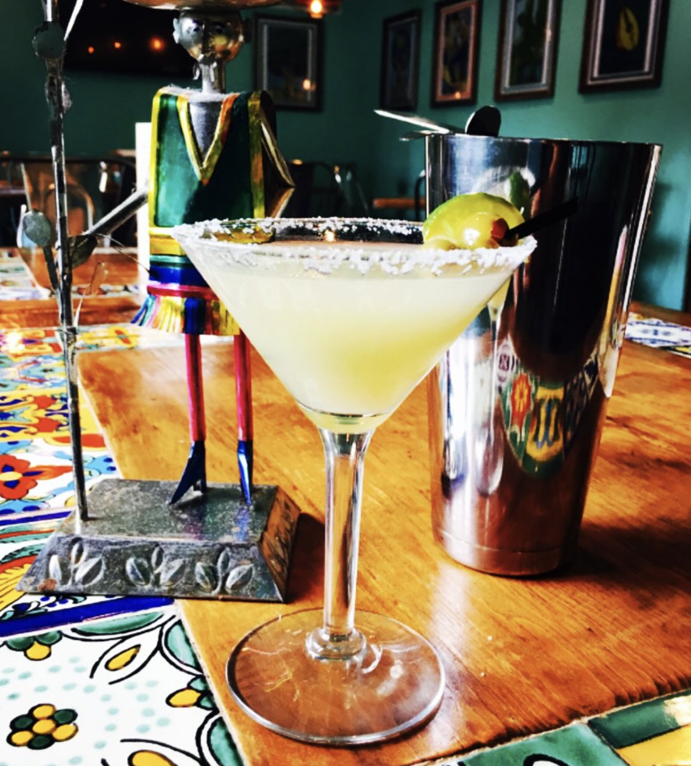 Salt-rimmed margarita, lime, shaker, and colorful figure on table.