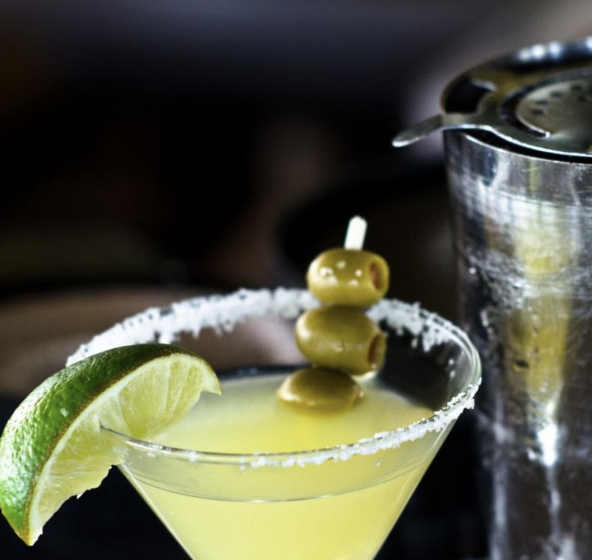 Yellow cocktail with salted rim, lime, olives, and shaker.