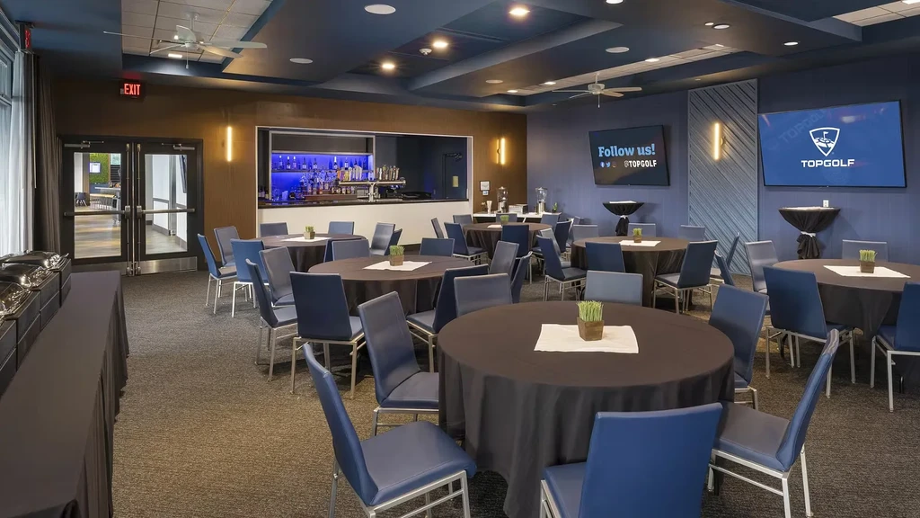Modern Topgolf event room with tables, blue chairs, buffet, and bar.