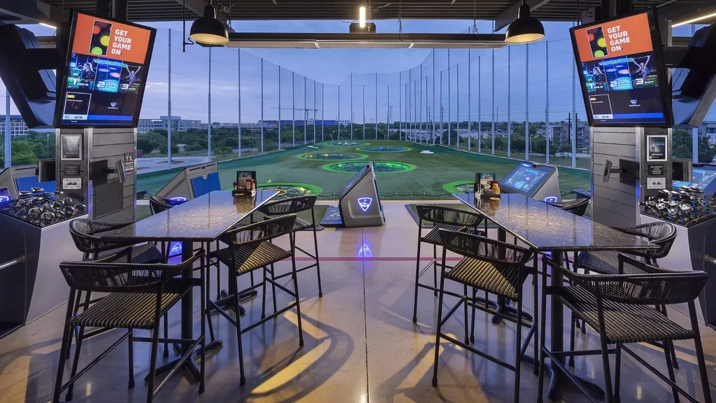 Empty Topgolf bays overlook an illuminated multi-level driving range at dusk.