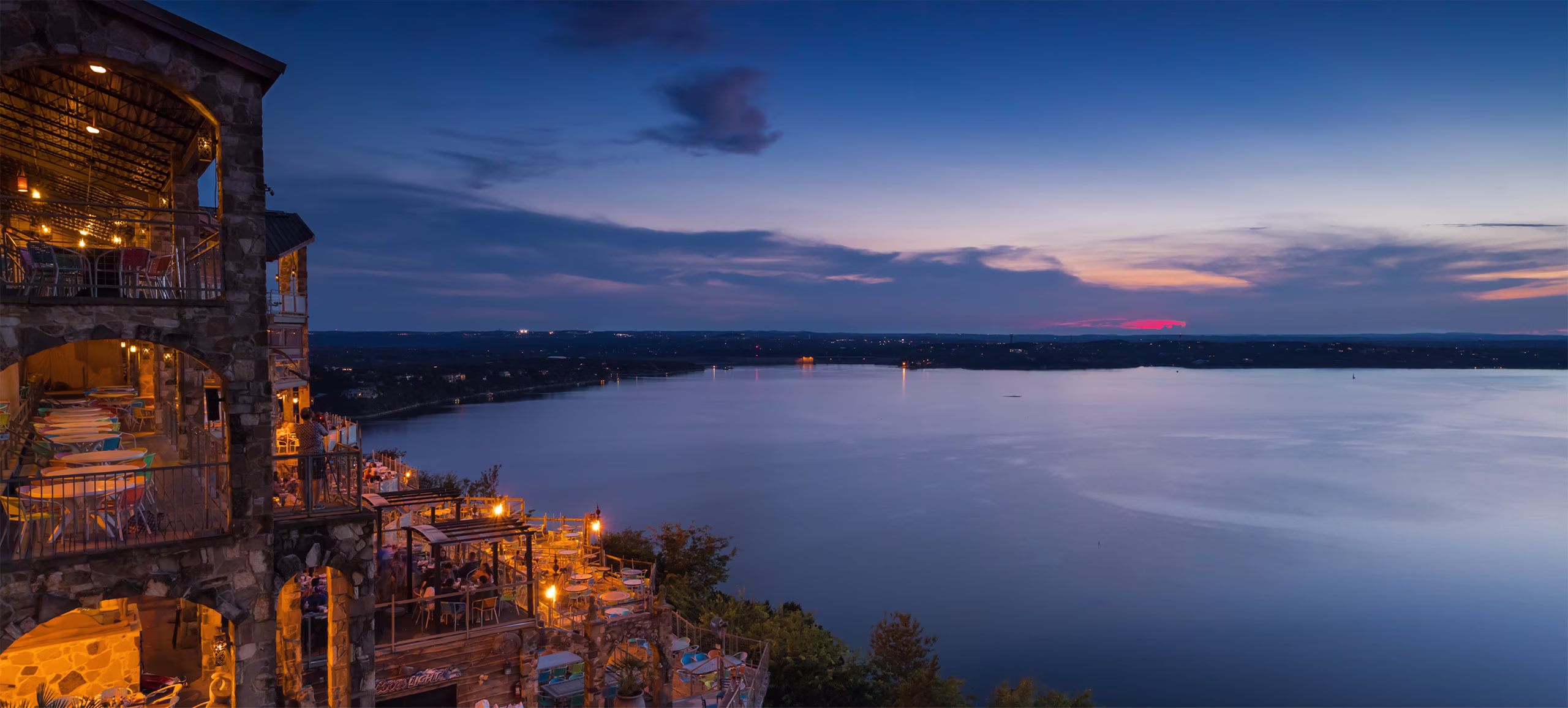 Illuminated stone restaurant overlooking a wide lake at twilight.