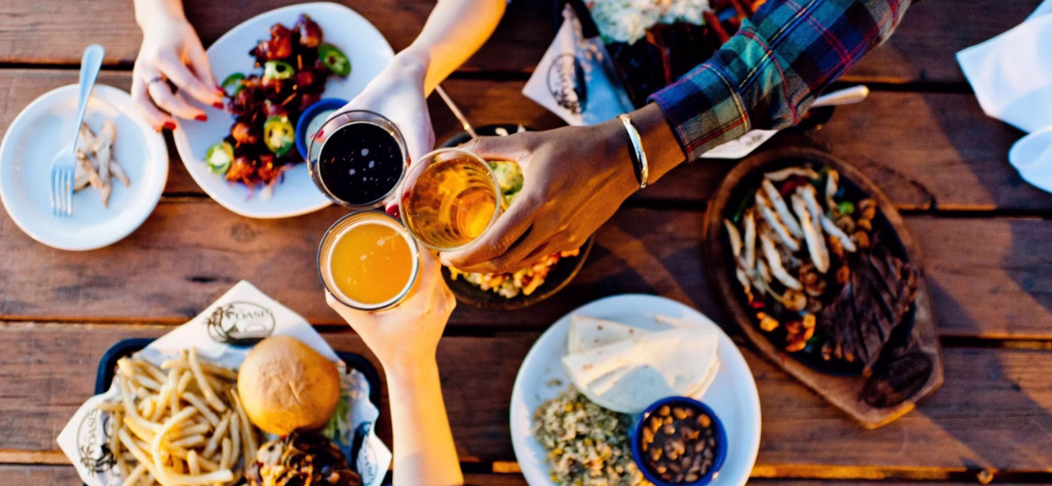 Three people clinking beer glasses over a table of food.
