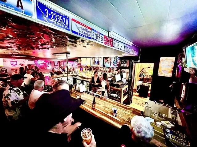 Interior of a busy bar with patrons, bartenders, and license plates.