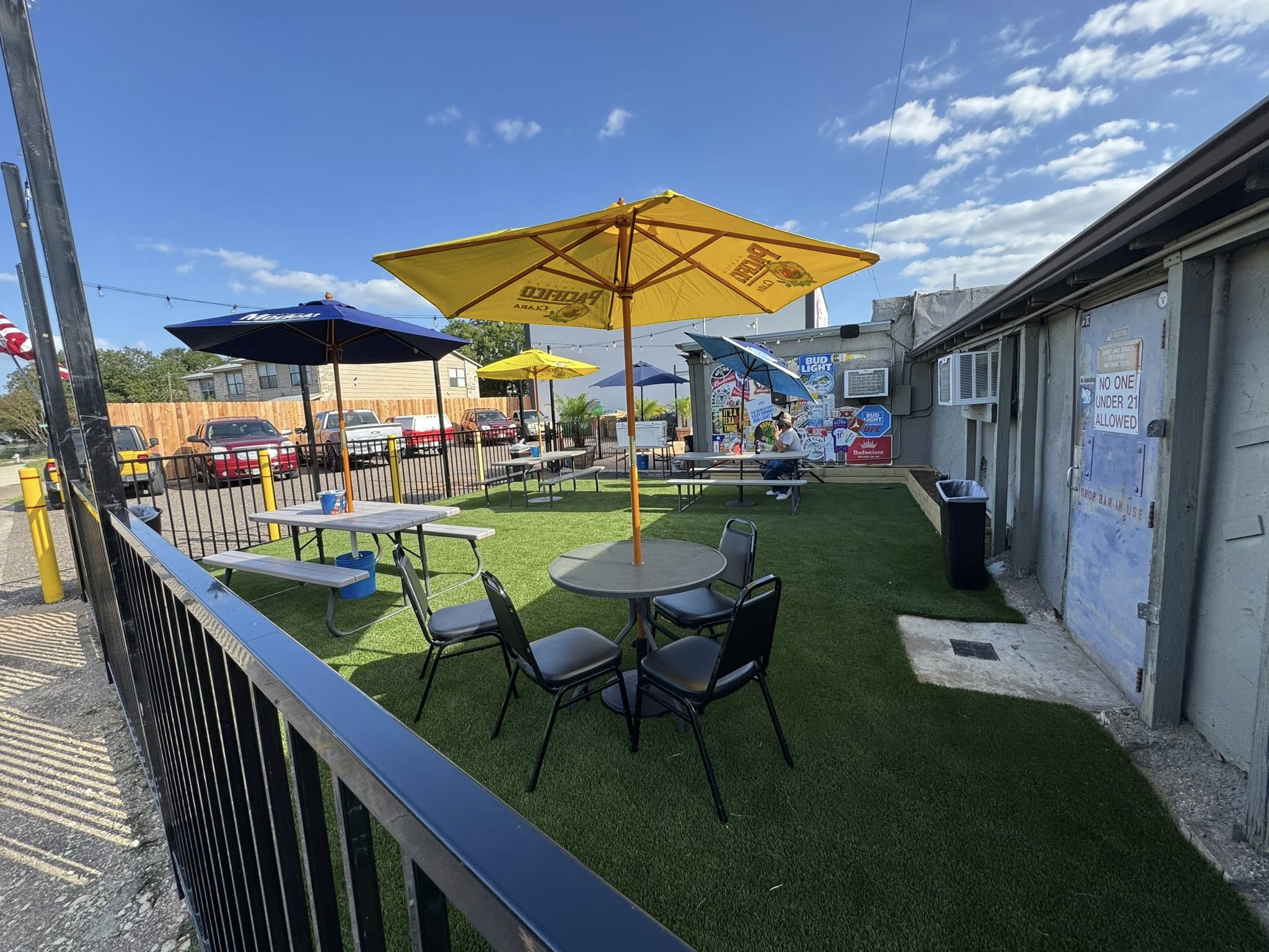 Outdoor patio with umbrellas, tables, chairs, and artificial turf under blue sky.