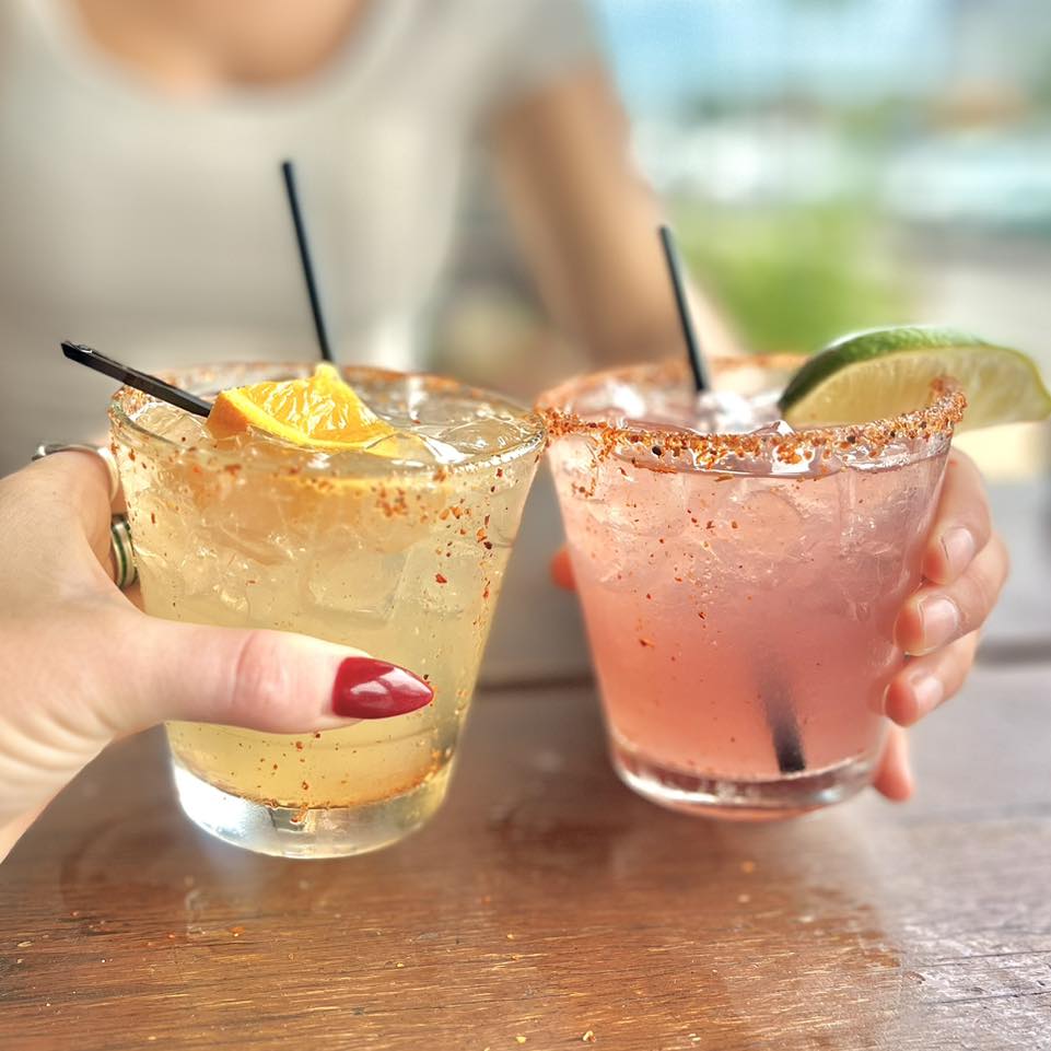 Two hands holding iced cocktails with chili rims, orange and lime.