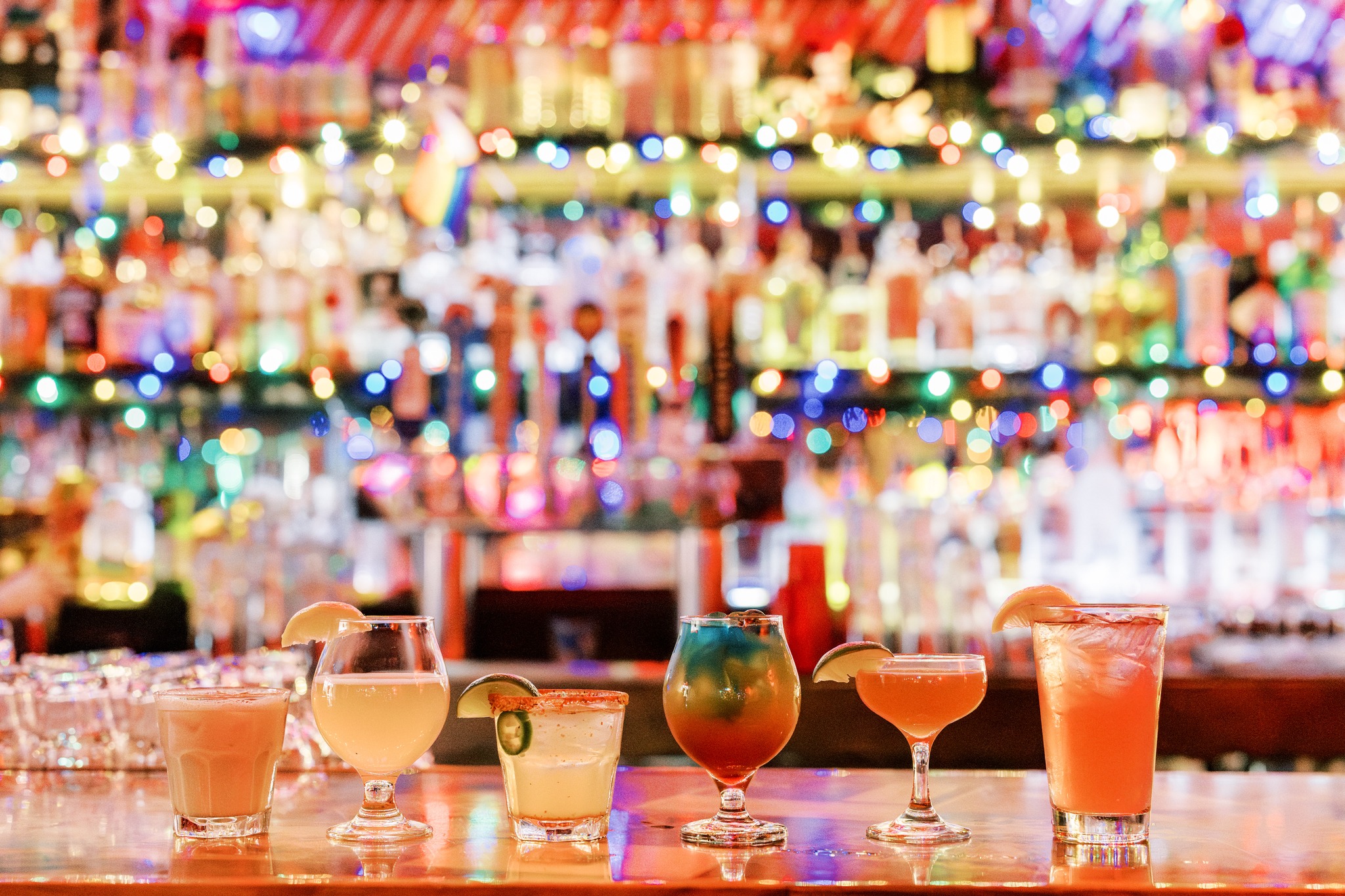 Six assorted cocktails on a bar with colorful lights background.