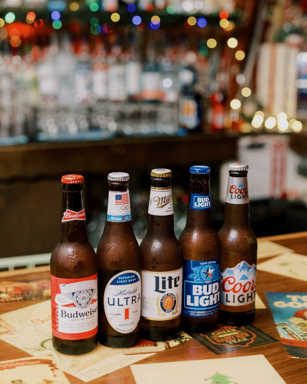 Five American beer bottles on a bar counter with blurred festive lights.