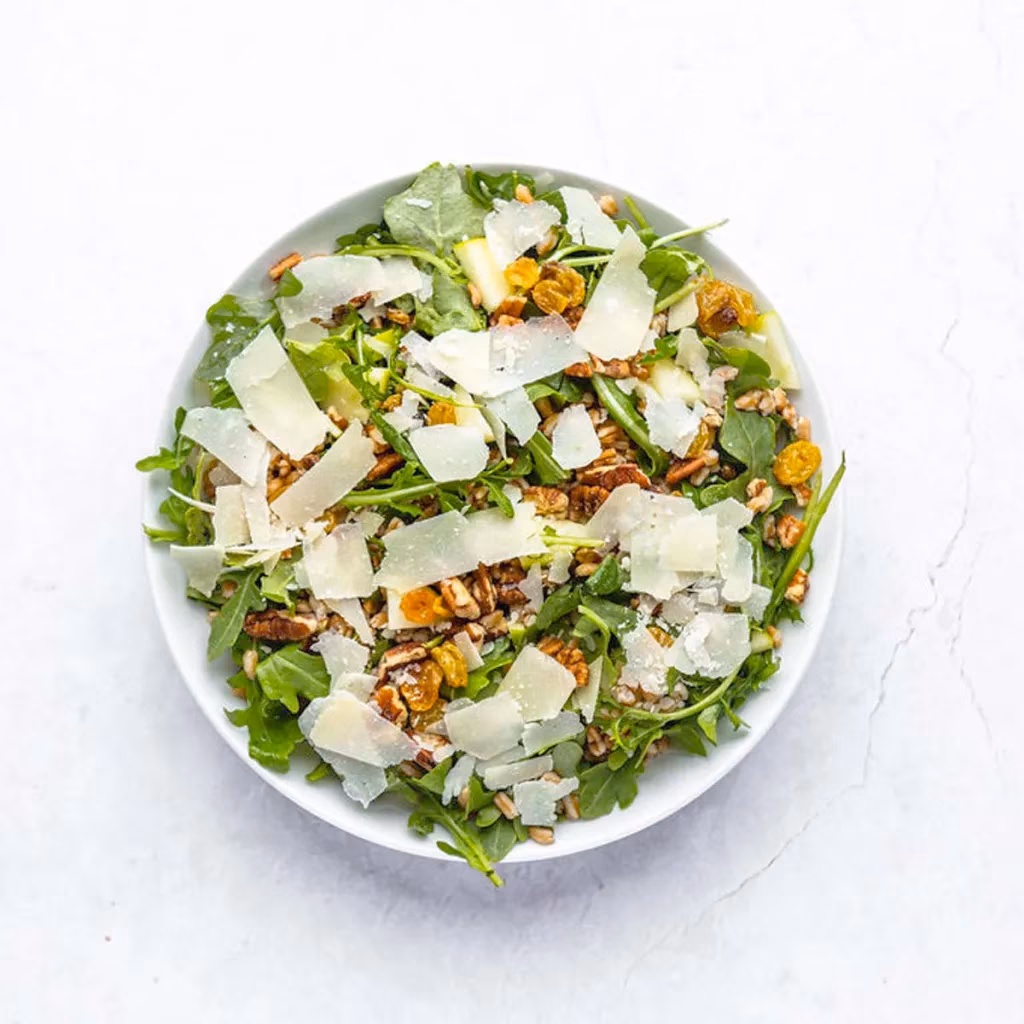 Arugula salad with shaved Parmesan, pecans, golden raisins in a white bowl.