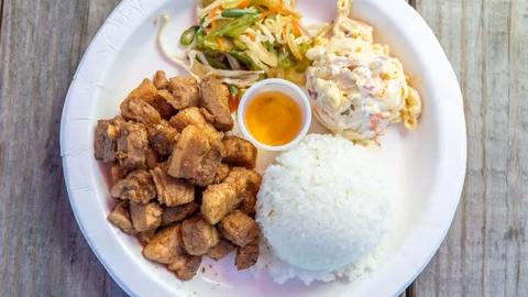 A white plate of pork chunks, rice, salad, macaroni, and dipping sauce.