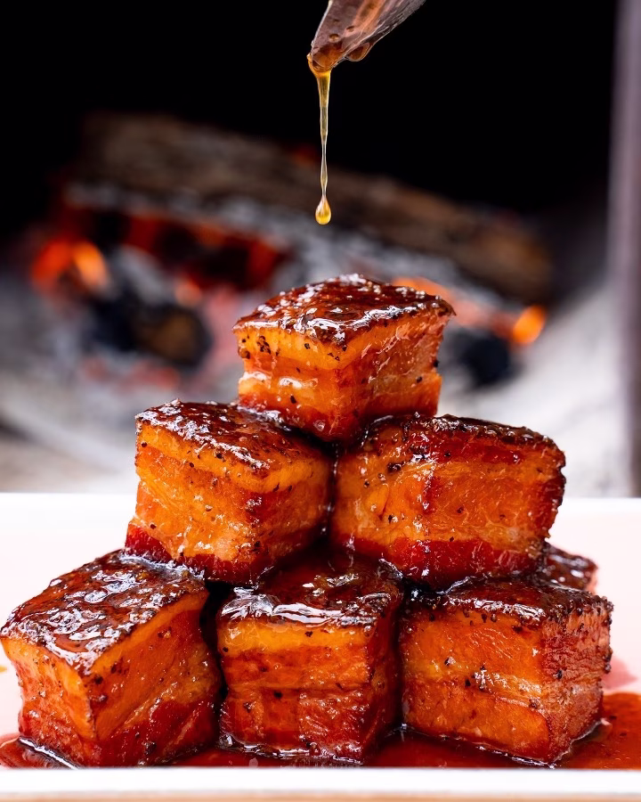 Stacked glazed meat cubes with dripping sauce and blurry fire background.