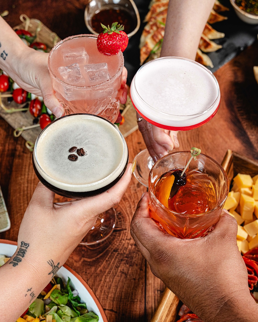 Four hands toasting with four different cocktails over a wooden table.