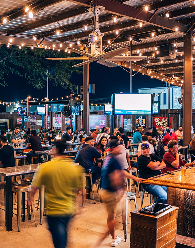 Crowded outdoor bar patio at night with string lights and a large fan.